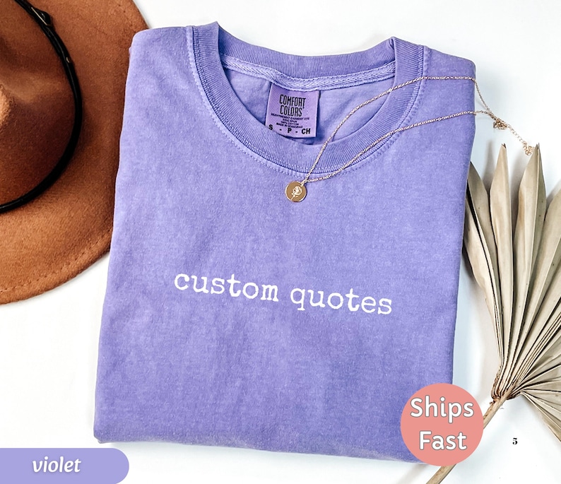 K&ouml;nnte beinhalten: Ein violettes T-Shirt mit den wei&szlig; gestickten Worten "custom quotes". Eine goldene Halskette mit Anh&auml;nger liegt auf dem Shirt. Ein brauner Hut und eine getrocknete Pflanze befinden sich im Hintergrund.