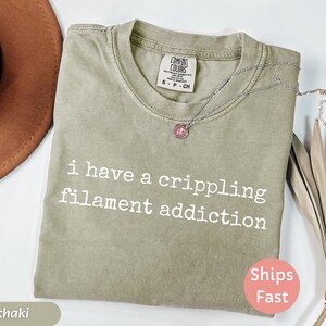 May include: Khaki-colored t-shirt with the text "i have a crippling filament addiction" in white. The shirt has a crew neck and a Comfort Colors tag. A silver necklace with a pendant is on the shirt. A "Ships Fast" sticker is visible.