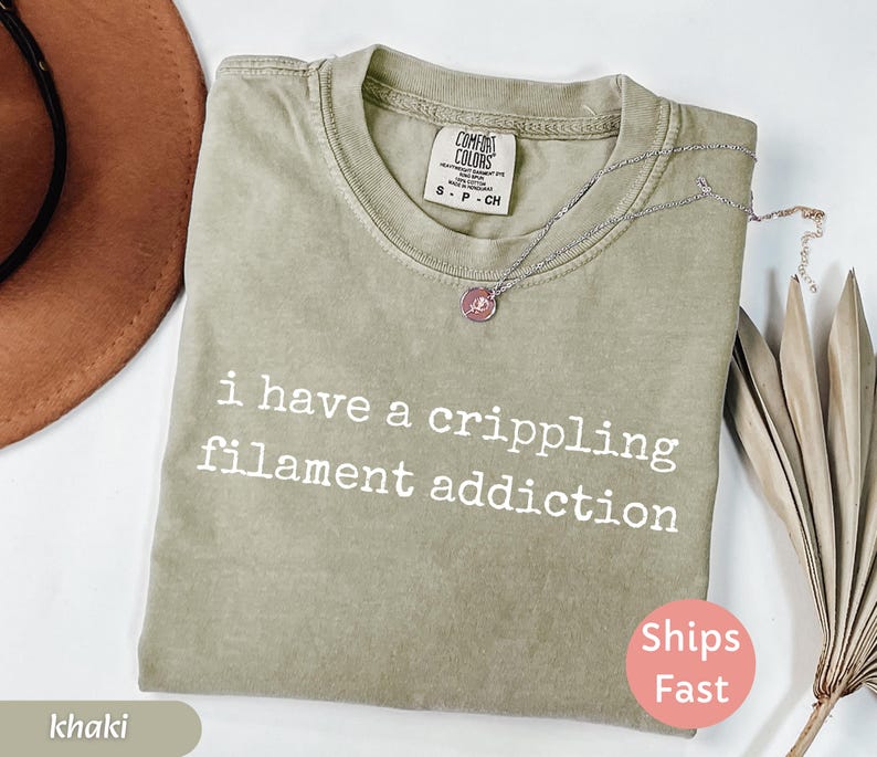 May include: Khaki-colored t-shirt with the text "i have a crippling filament addiction" in white. The shirt has a crew neck and a Comfort Colors tag. A silver necklace with a pendant is on the shirt. A "Ships Fast" sticker is visible.