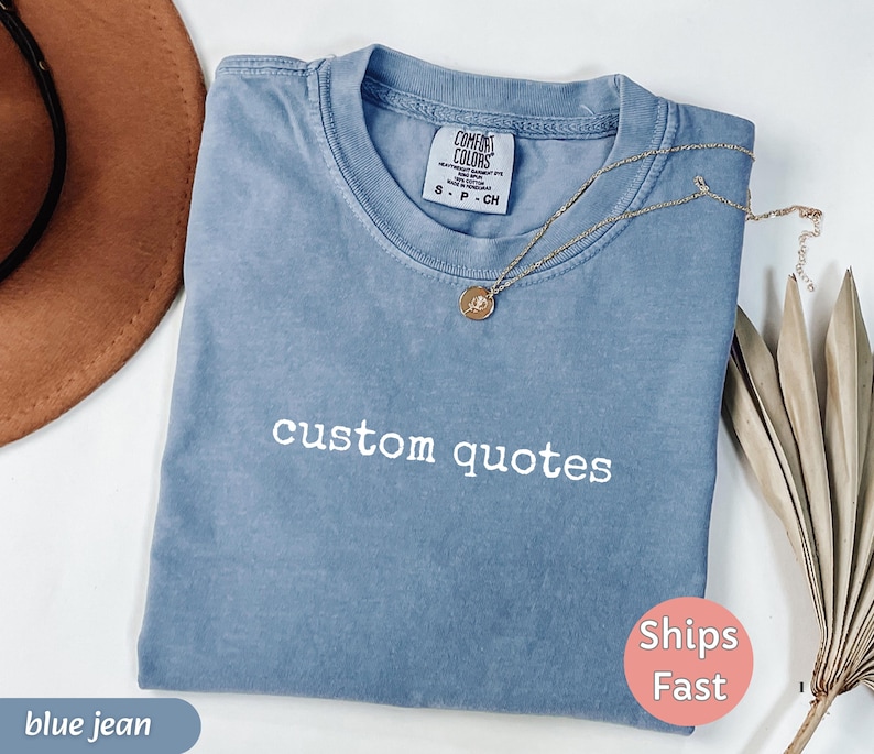 K&ouml;nnte beinhalten: Ein blaues Jeans-T-Shirt mit den Worten "custom quotes" in Wei&szlig; gestickt. Eine goldene Halskette mit Anh&auml;nger liegt auf dem Shirt. Das Shirt ist gefaltet und hat einen "Ships Fast"-Aufkleber.