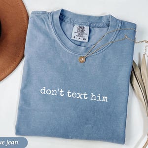 May include: A blue jean Comfort Colors t-shirt with the phrase "don't text him" printed in white. A gold necklace with a pendant is draped across the shirt. A brown hat and dried plant are in the background.