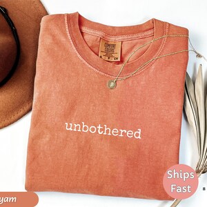 May include: A coral-colored t-shirt with the word "unbothered" embroidered in white. A gold necklace with a pendant rests on the shirt. A brown hat and dried plant are in the background. The shirt has a Comfort Colors tag.