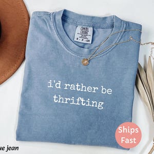 May include: A blue jean colored t-shirt with the phrase "i'd rather be thrifting" printed in white. A gold necklace with a pendant is draped across the shirt. The shirt is folded and has a "Ships Fast" sticker.
