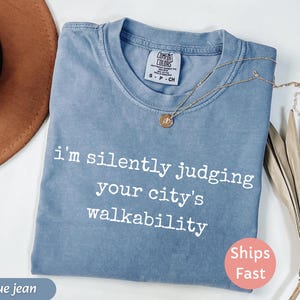 May include: A blue jean colored t-shirt with the text "I'm silently judging your city's walkability" in white. The shirt has a crew neck and short sleeves. A gold necklace with a pendant is draped across the shirt.