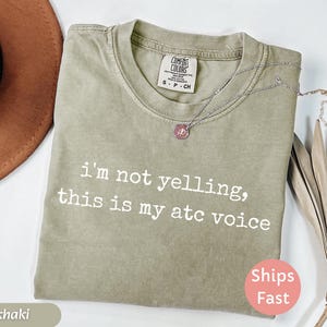 May include: Khaki-colored t-shirt with the text "I'm not yelling, this is my atc voice" in white. The shirt has a crew neck and a small tag near the collar. A silver necklace with a pendant is draped on the shirt.