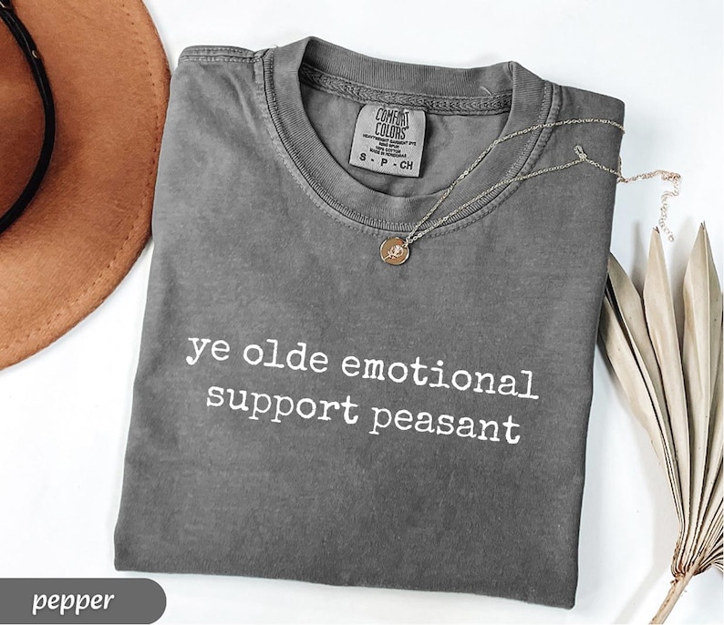 May include: Pepper-colored t-shirt with the text "ye olde emotional support peasant" in white. A silver necklace with a small pendant rests on the shirt. The shirt is folded and displayed on a white surface.
