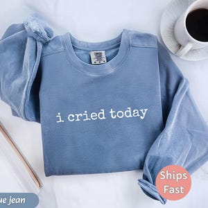 May include: A blue jean colored sweatshirt with the phrase "i cried today" printed in white. The sweatshirt is folded with a coffee cup and saucer in the background. The sweatshirt has a crew neck and long sleeves.