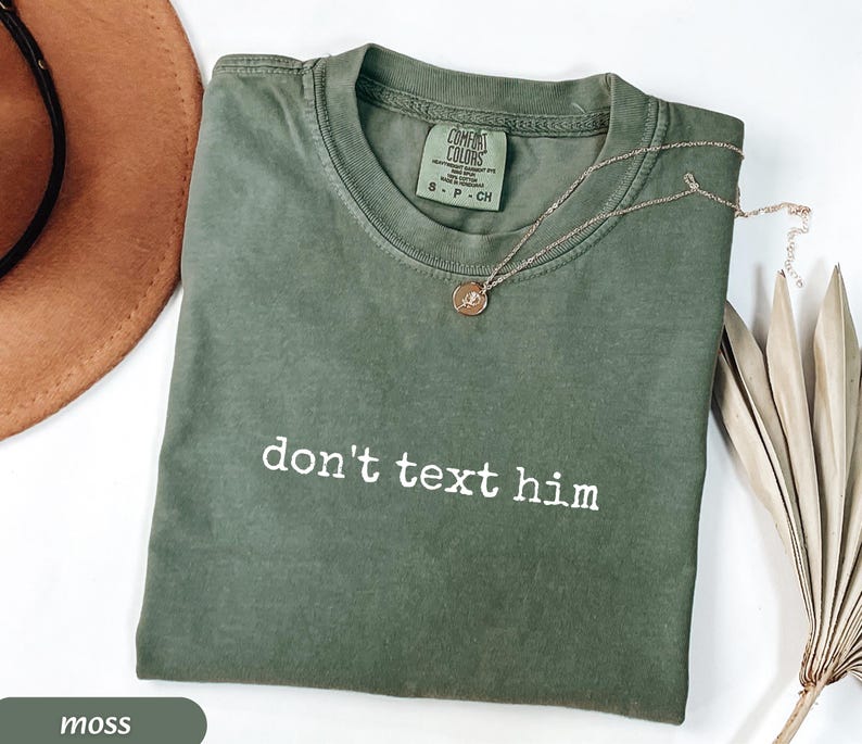 May include: Moss green t-shirt with the text "don't text him" in white. The shirt has a crew neck and short sleeves. A gold necklace with a pendant is draped across the shirt. The shirt is folded on a white surface.