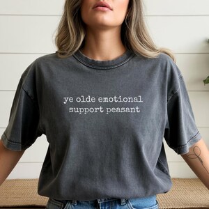 May include: Dark gray t-shirt with the text "ye olde emotional support peasant" in white. The shirt has a vintage, washed look and a crew neck. The short-sleeved tee is a casual style.