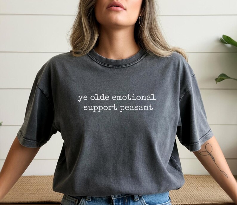 May include: Dark gray t-shirt with the text "ye olde emotional support peasant" in white. The shirt has a vintage, washed look and a crew neck. The short-sleeved tee is a casual style.