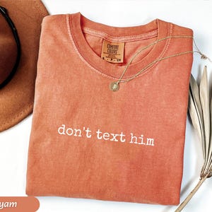 May include: A yam-colored Comfort Colors t-shirt with the phrase "don't text him" in white lettering. A gold necklace with a pendant rests on the shirt. A brown hat and dried plant are in the background.