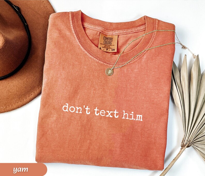 May include: A yam-colored Comfort Colors t-shirt with the phrase "don't text him" in white lettering. A gold necklace with a pendant rests on the shirt. A brown hat and dried plant are in the background.