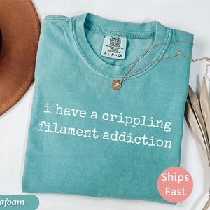 May include: Seafoam green t-shirt with the text "i have a crippling filament addiction" in white. The shirt has a crew neck and short sleeves. A gold necklace with a pendant is draped across the shirt.