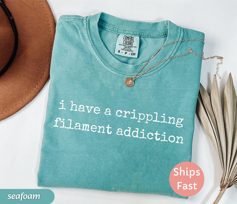 May include: Seafoam green t-shirt with the text "i have a crippling filament addiction" in white. The shirt has a crew neck and short sleeves. A gold necklace with a pendant is draped across the shirt.