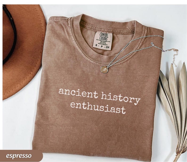Pu&ograve; includere: T-shirt color espresso con la scritta bianca "ancient history enthusiast". Una collana d'argento con un ciondolo &egrave; appoggiata sulla maglietta. Un cappello marrone e foglie secche sono sullo sfondo.