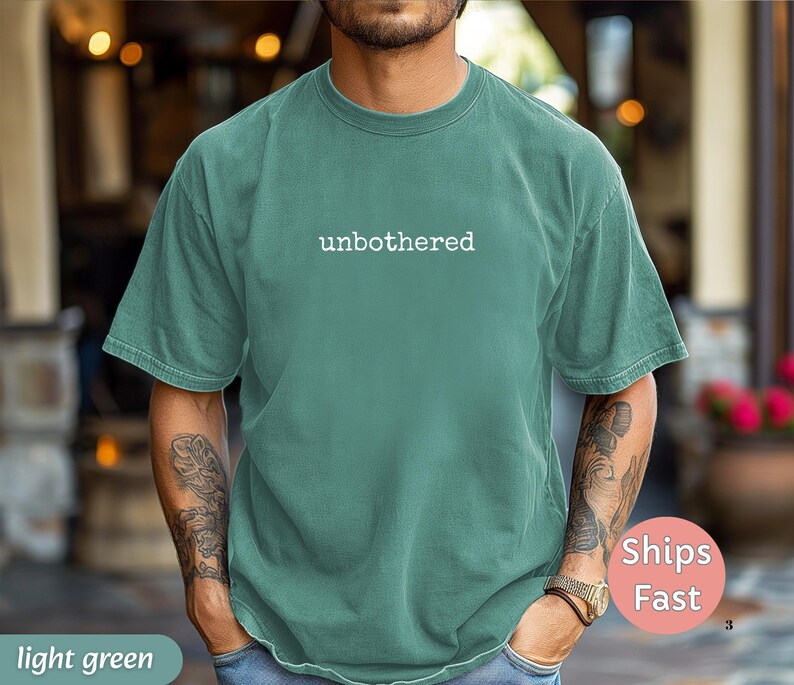 May include: Light green t-shirt with the word "unbothered" printed in white. The shirt has a crew neck and short sleeves. The model is wearing blue jeans and a watch.