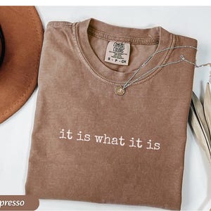 May include: Espresso-colored t-shirt with the phrase "it is what it is" in white lettering. A silver necklace with a small pendant rests on the shirt. The shirt is folded and next to a brown hat.