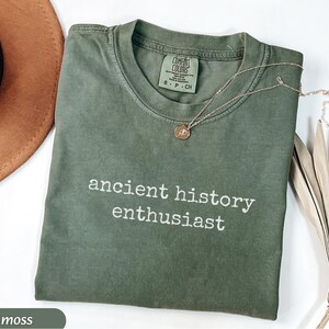 Pu&ograve; includere: Una maglietta verde muschio con le parole "ancient history enthusiast" in bianco. Una collana d'oro con un ciondolo poggia sulla maglietta. Un cappello marrone e una pianta secca sono sullo sfondo.