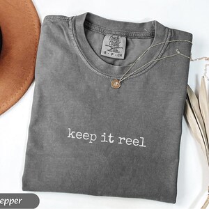 May include: A gray Comfort Colors t-shirt with the phrase "keep it reel" in white lettering. A silver necklace with a small pendant rests on the shirt. A brown hat and dried palm leaves are in the background.