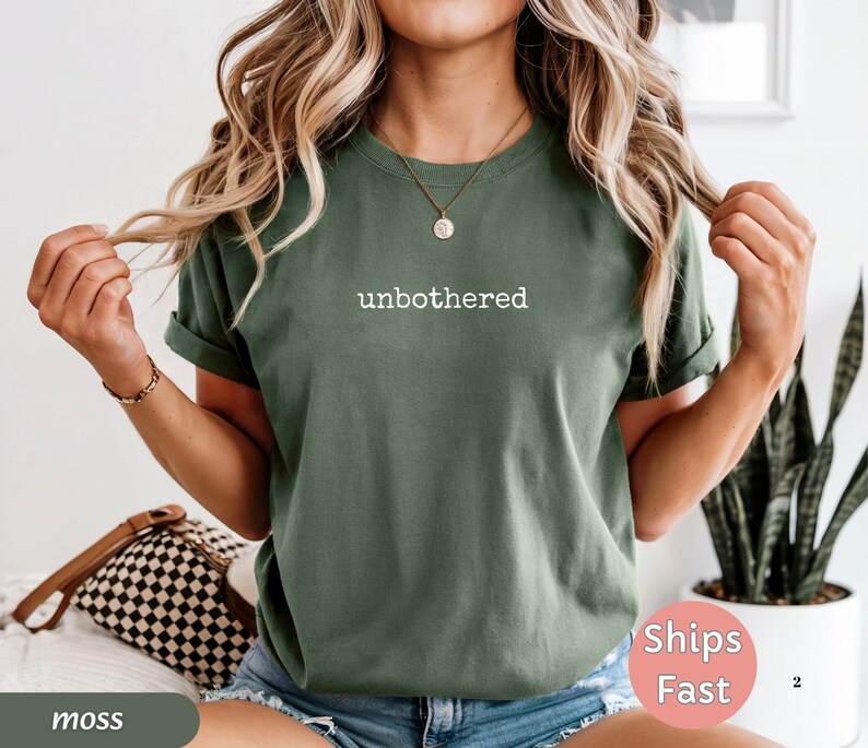 May include: Moss green t-shirt with the word "unbothered" printed in white. The shirt has rolled-up sleeves and is paired with blue denim shorts. A brown and black checkered purse is visible.