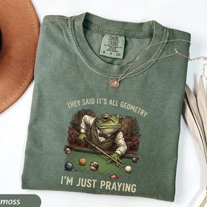 May include: Moss green t-shirt with a graphic of a frog playing pool, wearing a suit and bow tie. The text reads "THEY SAID IT'S ALL GEOMETRY, I'M JUST PRAYING." A silver necklace is draped across the shirt.