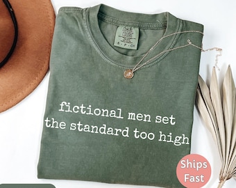 May include: A moss green t-shirt with the text "fictional men set the standard too high" in white. The shirt has a crew neck and a small tag. A gold necklace with a pendant is on the shirt.