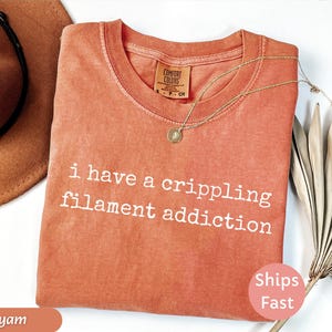 May include: A coral-colored t-shirt with the text "i have a crippling filament addiction" in white. A gold necklace with a pendant rests on the shirt. A brown hat and dried palm leaves are also in the image.