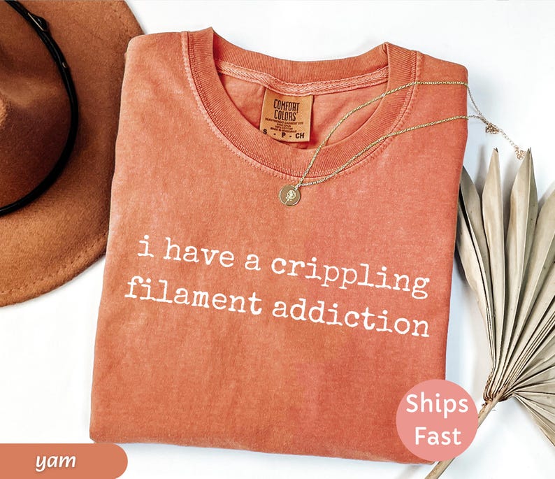 May include: A coral-colored t-shirt with the text "i have a crippling filament addiction" in white. A gold necklace with a pendant rests on the shirt. A brown hat and dried palm leaves are also in the image.