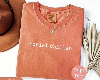 Puede incluir: Una camiseta de color coral con las palabras "serial chiller" en letras blancas. Un collar de oro con un colgante descansa sobre la camisa. Un sombrero marrón y hojas de palma secas están en el fondo.