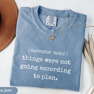 May include: A blue jean colored t-shirt with the text "(narrator voice) things were not going according to plan." A gold necklace with a pendant is draped across the shirt. The shirt is folded and displayed on a white surface.