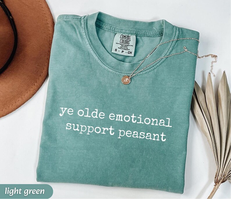 May include: A light green t-shirt with the text "ye olde emotional support peasant" in white. A gold necklace with a pendant rests on the shirt. The shirt is folded and has a tag that says "Comfort Colors".