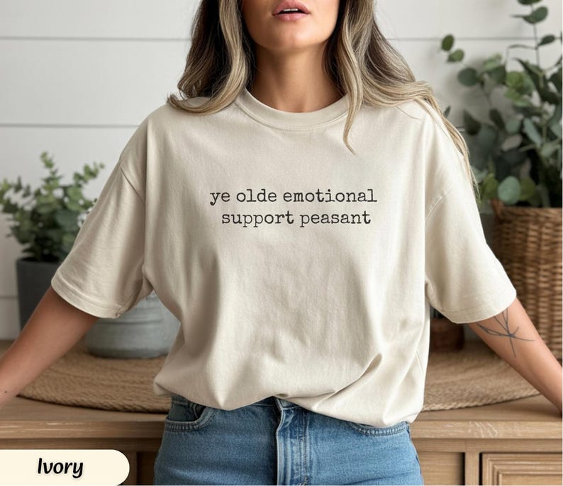 May include: Ivory-colored t-shirt with the text "ye olde emotional support peasant" in black. The shirt is a relaxed fit and is paired with blue jeans. The overall style is casual and comfortable.