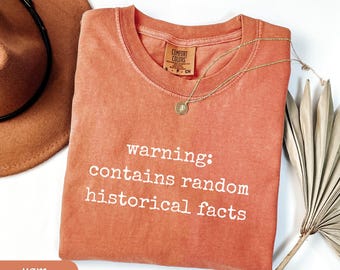 May include: A coral-colored t-shirt with the text "warning: contains random historical facts" in white. The shirt has a crew neck and a small tag. A gold necklace and a brown hat are also visible.