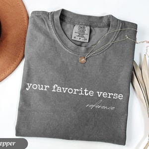 May include: A gray Comfort Colors t-shirt with the text "your favorite verse reference" in white. A gold necklace with a pendant rests on the shirt. A brown hat and dried plant are in the background.