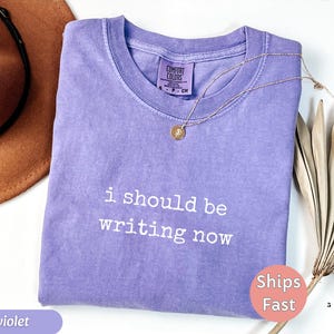May include: Violet-colored t-shirt with the text "i should be writing now" in white. The shirt has a crew neck and short sleeves. A gold necklace with a pendant is draped on the shirt. A brown hat and dried plant are in the background.