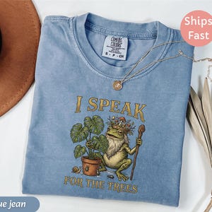 May include: Blue jean Comfort Colors t-shirt with the phrase "I Speak For The Trees" and a graphic of a frog with a plant. A gold necklace with a pendant is on the shirt. The shirt is folded.