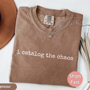 May include: Espresso-colored t-shirt with the phrase "i catalog the chaos" in white. The shirt has a crew neck and a Comfort Colors tag. A silver necklace with a pendant is draped on the shirt. A brown hat and dried leaves are in the background.