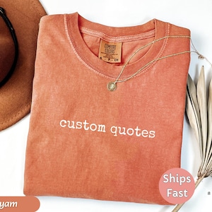K&ouml;nnte beinhalten: Ein korallenfarbenes T-Shirt mit den Worten "custom quotes" in Wei&szlig; gestickt. Eine goldene Halskette mit Anh&auml;nger liegt auf dem Shirt. Ein brauner Hut und getrocknete Palmbl&auml;tter sind im Hintergrund. Das Shirt hat ein "Comfort Colors"-Etikett.