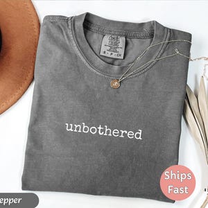 May include: A gray t-shirt with the word "unbothered" in white lettering. A gold necklace with a pendant rests on the shirt. A brown hat and dried leaves are in the background.