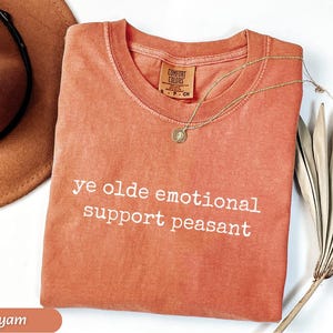 May include: A yam-colored Comfort Colors t-shirt with the phrase "ye olde emotional support peasant" in white lettering. A gold necklace with a pendant and a brown hat are also visible.
