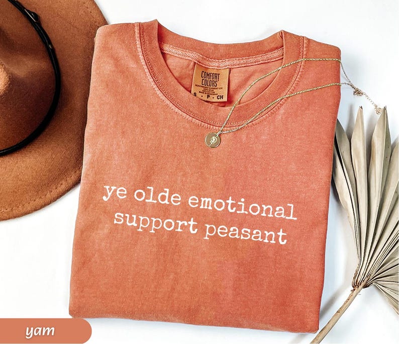 May include: A yam-colored Comfort Colors t-shirt with the phrase "ye olde emotional support peasant" in white lettering. A gold necklace with a pendant and a brown hat are also visible.