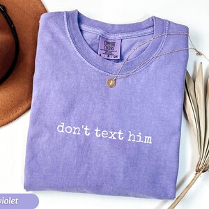 May include: A violet Comfort Colors t-shirt with the phrase "don't text him" in white lettering. A gold necklace with a pendant rests on the shirt. A brown hat and dried palm leaves are in the background.