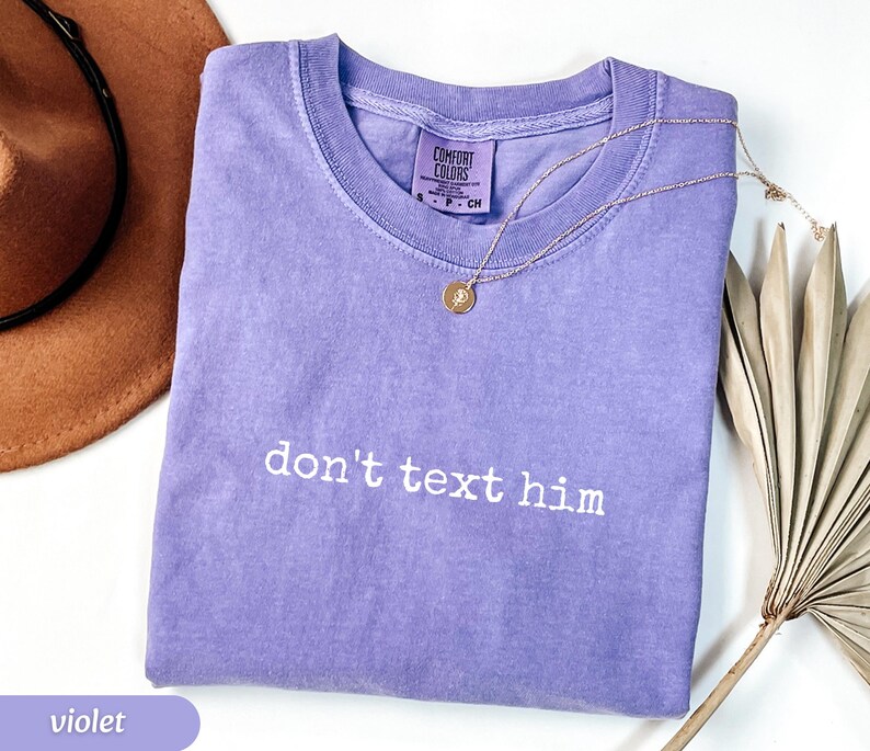 May include: A violet Comfort Colors t-shirt with the phrase "don't text him" in white lettering. A gold necklace with a pendant rests on the shirt. A brown hat and dried palm leaves are in the background.