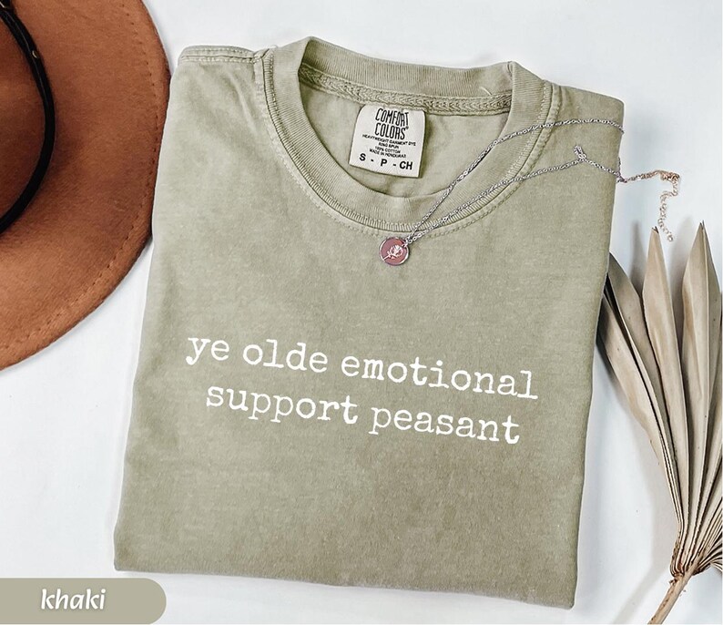May include: Khaki-colored t-shirt with the text "ye olde emotional support peasant" in white. A silver necklace with a pink pendant rests on the shirt. The shirt is folded and displayed with a brown hat and dried leaves.