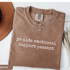 May include: Espresso-colored t-shirt with the text "ye olde emotional support peasant" in white. A silver necklace with a small pendant rests on the shirt. The shirt is folded, and a brown hat is visible in the background.