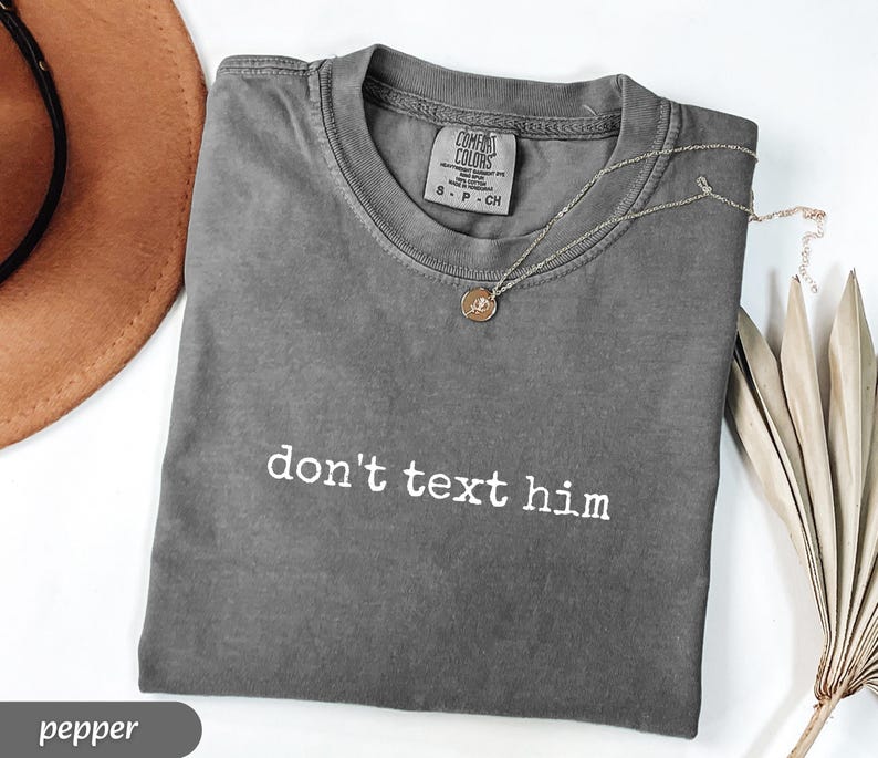 May include: A gray t-shirt with the phrase "don't text him" in white lettering. A gold necklace with a pendant rests on the shirt. A brown hat and dried plant are in the background.
