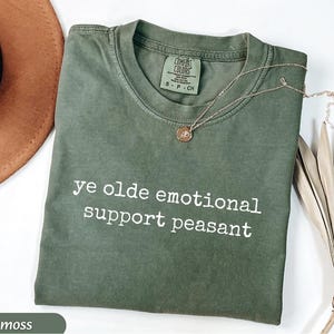 May include: A moss green t-shirt with the text "ye olde emotional support peasant" in white. A gold necklace with a pendant rests on the shirt. The shirt is folded on a white surface.