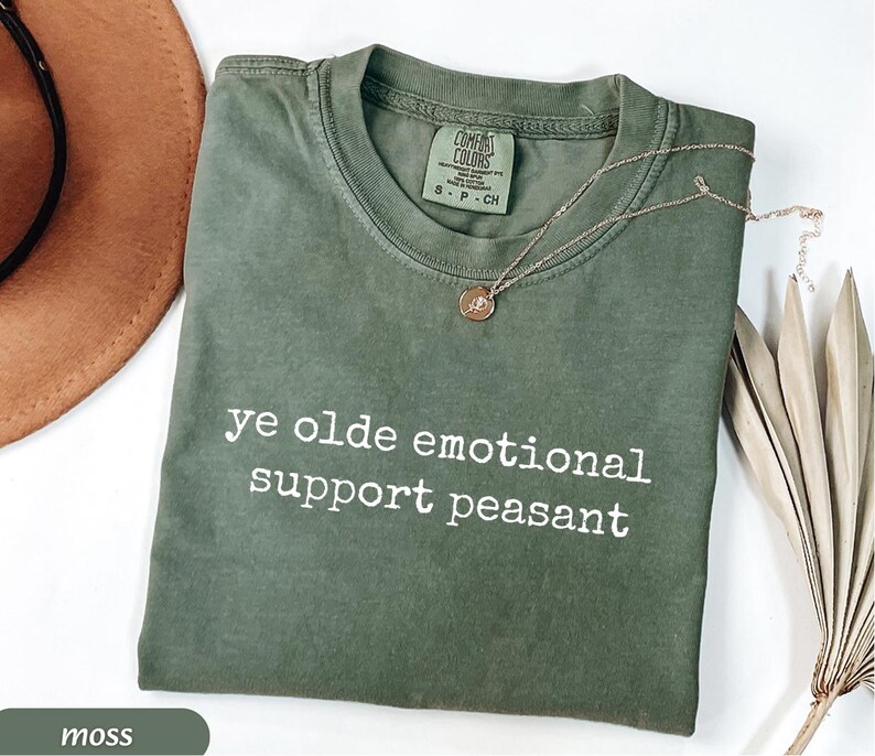May include: A moss green t-shirt with the text "ye olde emotional support peasant" in white. A gold necklace with a pendant rests on the shirt. The shirt is folded on a white surface.