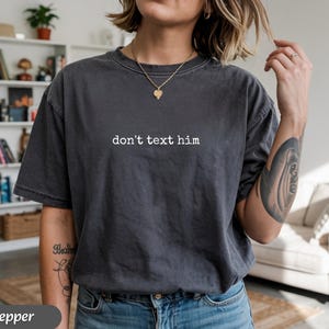 May include: Dark gray t-shirt with the phrase "don't text him" printed in white. The shirt has a crew neck and short sleeves. The word "pepper" is printed in a dark gray banner at the bottom of the image.