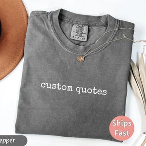K&ouml;nnte beinhalten: Ein graues T-Shirt mit den wei&szlig;en Worten "custom quotes". Eine goldene Halskette mit Anh&auml;nger liegt auf dem Shirt. Das Shirt ist gefaltet und hat ein Etikett mit der Aufschrift "Comfort Colors". Ein brauner Hut ist im Hintergrund.